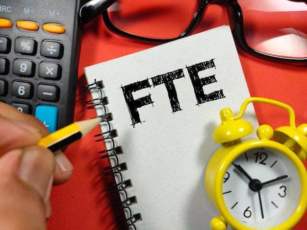 Text FTE Full Time Equivalent writing on notebook with hand holding pencilclockcalculator and glasses on red background