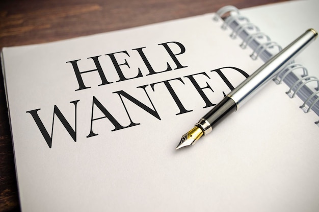 Text HELP WANTED on paper notepad and pen on wooden background