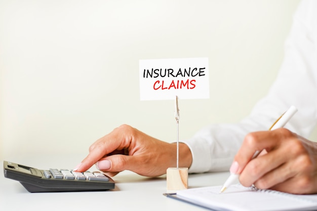 Photo the text insurance claims written on a white paper card clamped in a paper holder. in the background woman's hands writing with pen on paper and touch calculator