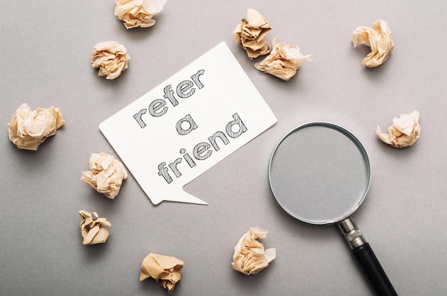 Text refer a friend on a white speech bubble on a gray background surrounded by crumpled notes and a magnifying glass