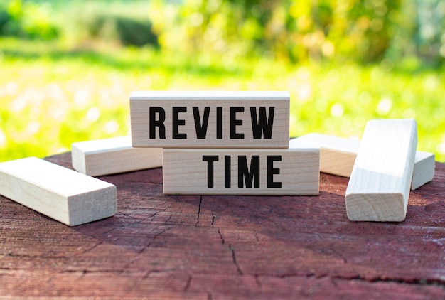 The text REVIEW TIME is written on a wooden cubes lying on an old tree stump against a blurred garden background