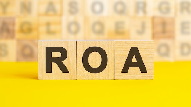 The text ROA is written on wooden cubes on a bright yellow surface. In the background are rows of cubes with different letters. Business concept. ROA - short for return on assets