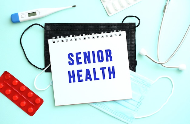 The text SENIOR HEALTH is written on a white sheet of notepad that lies on a blue background next to a thermometer and medical face masks