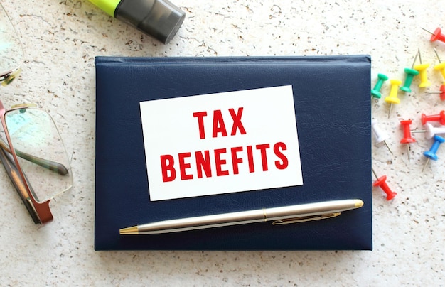 Text TAX BENEFITS on a business card lying on a blue notebook next to the glasses and stationery