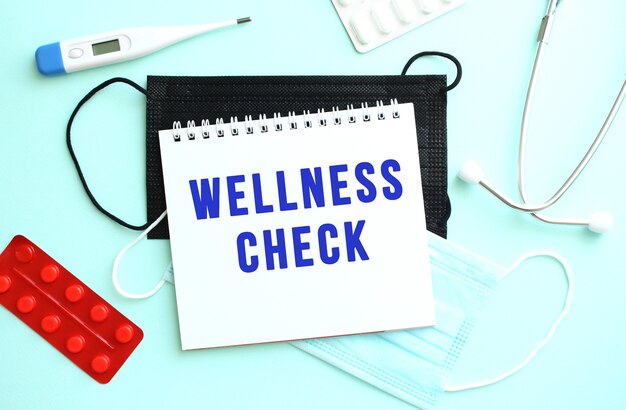 The text WELLNESS CHECK is written in a notebook that lies on a blue background next to a thermometer and medical masks.