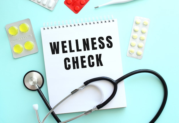 The text WELLNESS CHECK is written on a white notepad that lies next to the stethoscope and pills on a blue background