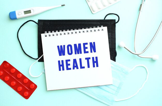 The text WOMEN HEALTH is written on a white sheet of notepad that lies on a blue background next to a thermometer and medical face masks. Medical concept