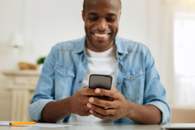 Texting. Attractive happy dark-haired afro-american man smiling and writing a message on his phone while sitting at the table