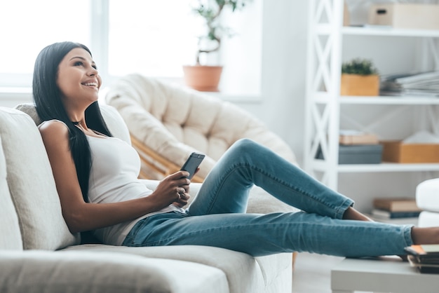 Texting to boyfriend. Attractive young woman holding mobile phone and looking away while relaxing on the couch at home