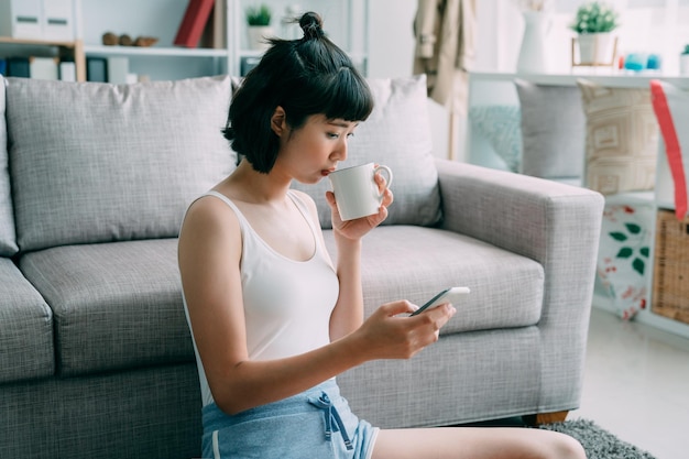 Texting to boyfriend. beautiful young asian chinese woman in shorts holding cup of coffee and using smartphone while relax on floor at home living room. attractive girl text sms online on cellphone