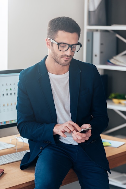 Texting business message. Confident young man holding smart phone and looking at it while leaning at the office desk