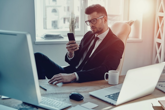 Texting to client. Handsome young man in formalwear using his smart phone while sitting in the office