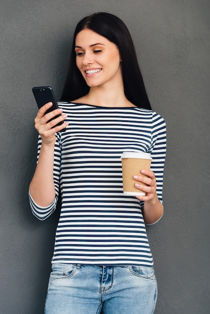 Texting to friend. Attractive young smiling woman holding coffee cup and looking at her smart phone while standing