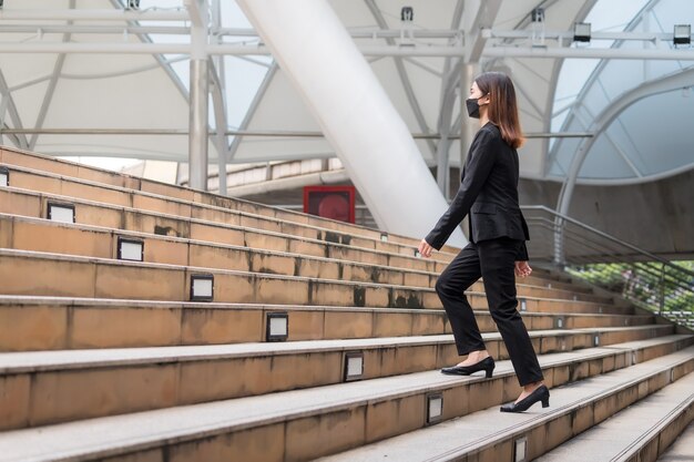 A Thai Asian woman wearing a black suit, trousers and a mask. She was walking up the stairs. In the concept of travel and advancement of a business woman or office worker.