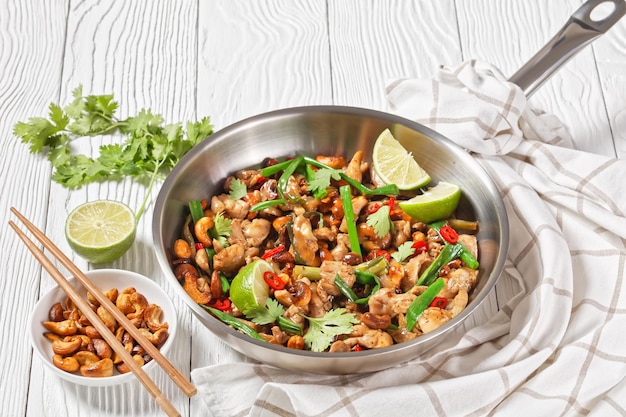 Thai cashew chicken stir fry in a pan on a wooden table with chopsticks and ingredients horizontal view from above closeup