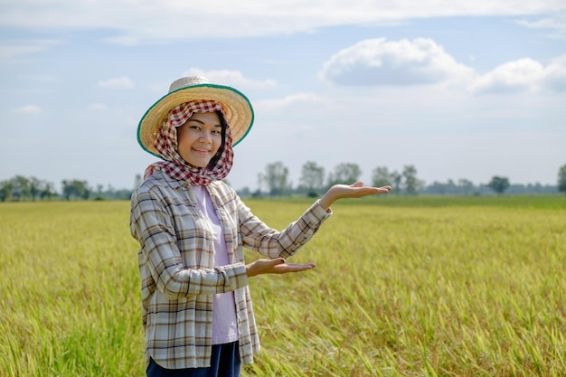 A Thai female farmer wearing a traditional shirt and hat is standing and posing holding product in the middle of the field