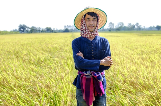 Thai male farmer in traditional dress and headdress standing with arms crossed smiling in the middle of the rice field