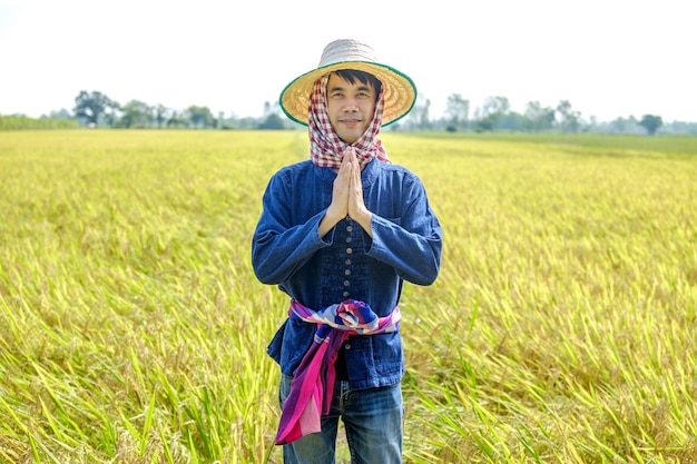 A Thai male farmer wearing a traditional blue shirt and hat is standing and posing thankyou in the middle of the field