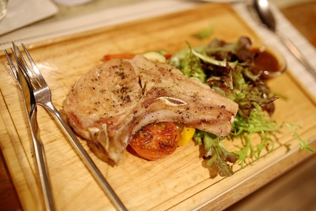 Thai style pork steak With roasted tomatoes side vegetables and spicy sauce blurred on a wooden plate looks delicious There is a knife and fork on the left Very shallow focus