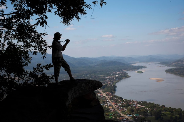 Thai woman travel visit and posing take photo on ridge stone of cliffs at Wat Pha Tak Suea temple with view of landscape of Nongkhai city and loas and Mekong river in Nong Khai Thailand