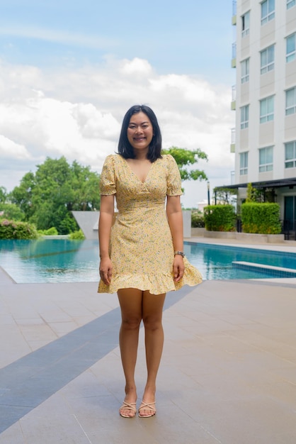 Thai Woman wearing yellow floral dress outdoors with swimming pool on background and greenery