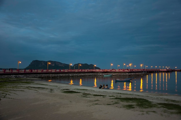 Thai women and friend sit relax playing mobile smart phone on sand beach with people playing skateboard Saphan saranwithi red bridge in night time on September 6 2021 in Prachuap Khiri Khan Thailand