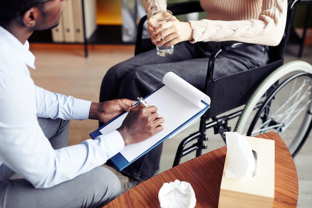 Therapist talking to young woman in wheelchair asking for her concerns and feelings and taking notes