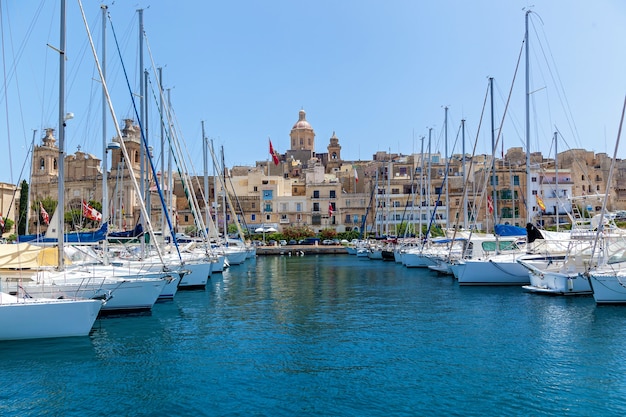 There are a lot of moored yachts in the harbor on the background of Maltese city with old buildings and palm trees on a summer sunny day.