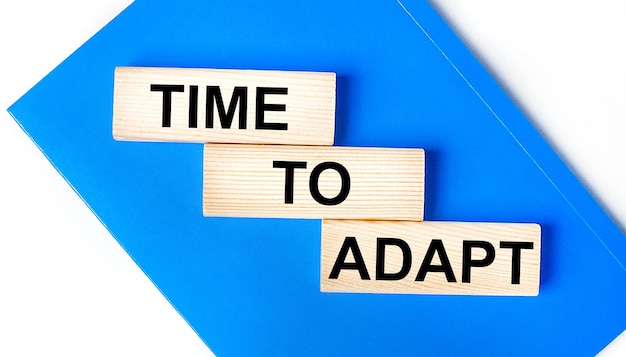 Photo there is a blue notebook on a light gray background above are three wooden blocks with the words time to adapt