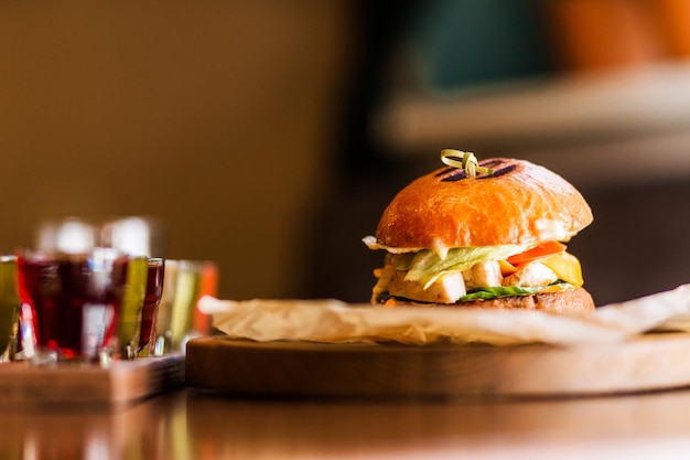 There is a burger with chicken cheese sauce tomato and greens on a table on a wooden platform Next to the burger is a set of alcoholic liqueurs Horizontal photo