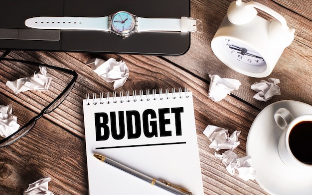 Photo there is a cup of coffee on a wooden table, a clock, glasses and a notebook with the word budget. business concept