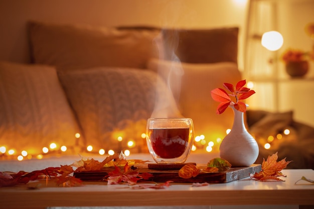 Thermo glass with hot tea in dark night room with autumnal leaves