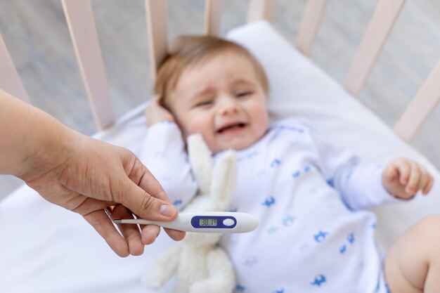 Thermometer in focus in mom's hands with a high temperature of 38 degrees on the background of a sick crying baby boy in a crib at home on white bedding medicine and pediatrics