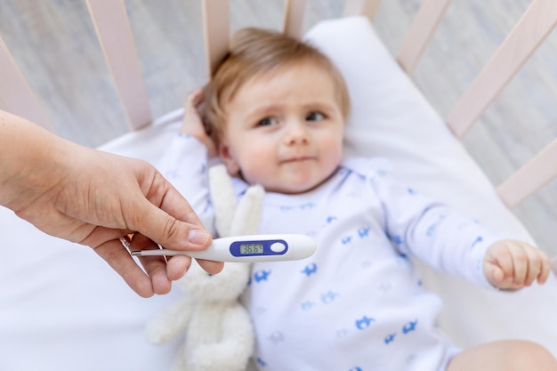 Thermometer in focus in mom's hands with a normal temperature of 366 degrees on the background of a healthy baby boy in a crib at home on white bedding medicine and pediatrics