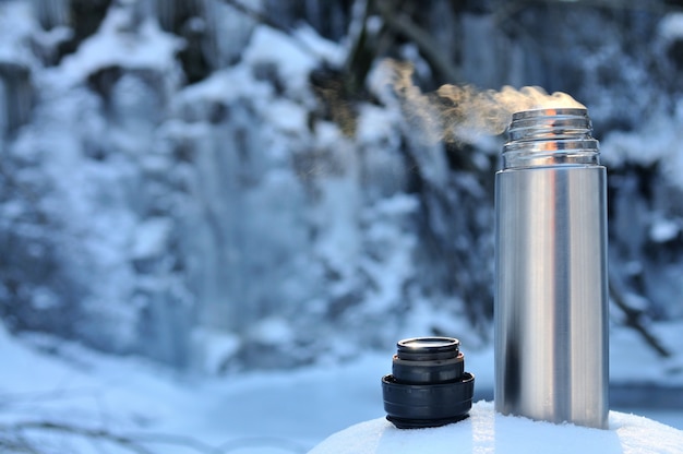 Thermos with a hot drink on mountain waterfall