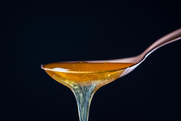 Thick honey dripping from the one metal spoon on black background close up Honey flowing from spoon