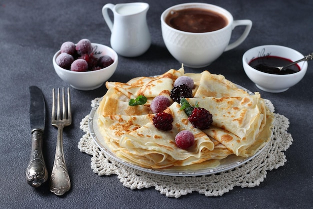 Thin pancakes with wheat flour, eggs and kefir, served with berries, jam and cup of cofee on dark gray table.