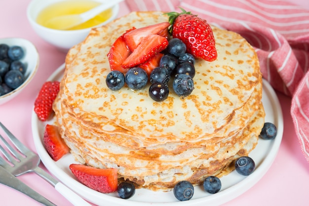 Thin traditional Russian pancakes on a trendy pink surface with berries and honey. Close up