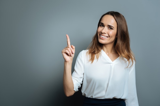 Thing to remember. Joyful positive happy woman smiling and pointing up with her finger while standing against grey background