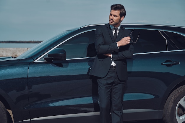 Thinking about... Handsome young businessman keeping arms crossed and smiling while standing near his car outdoors