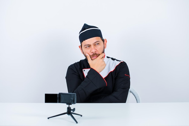 Thinking blogger chef is putting his hand on chin by sitting in front of his mini camera on white background