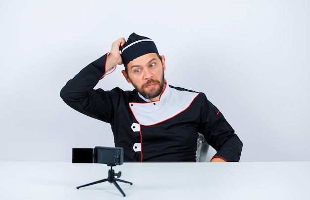 Thinking blogger chef is putting his hand on head by sitting in front of his mini camera on white background