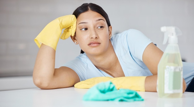 Thinking cleaner and woman at table in home tired of spring cleaning the kitchen interior Housekeeper girl with domestic gloves and detergent unhappy frustrated and moody with tidy job