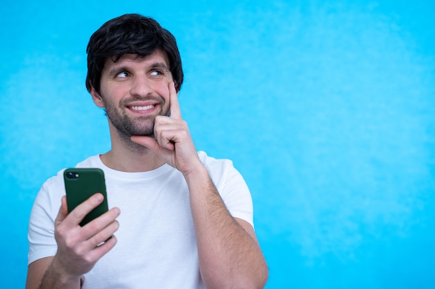 Thinking dreaming man using mobile phone posing over blue wall surface