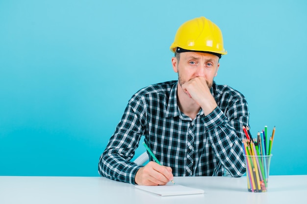 Thinking engineer man is looking at camera by holding hand on chin on blue background