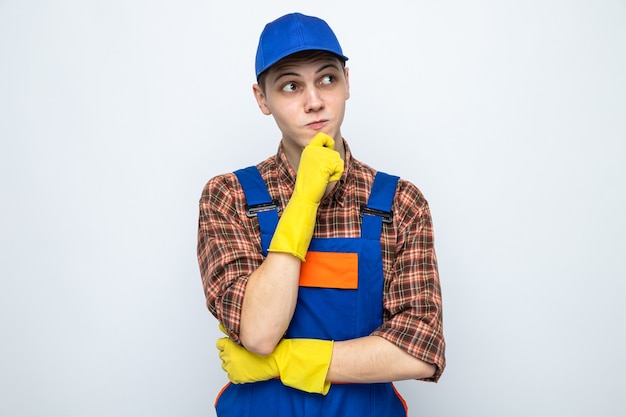 Thinking grabbed chin young cleaning guy wearing uniform and cap with gloves 