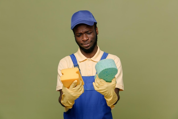 Thinking holding cleaning sponges young africanamerican cleaner male in uniform with gloves isolated on green background