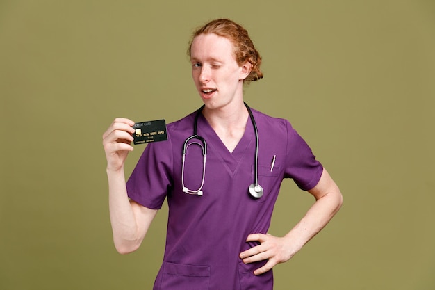 Thinking putting hand on hips young male doctor wearing uniform with stethoscope holding card isolated on green background