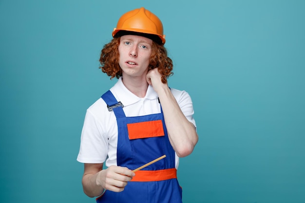 Thinking young builder man in uniform holding pencil isolated on blue background