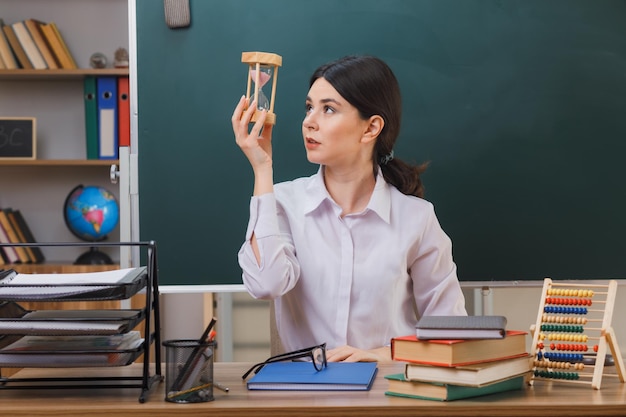 thinking young female teacher holding and looking at sand watch sitting at desk with school tools in classroom